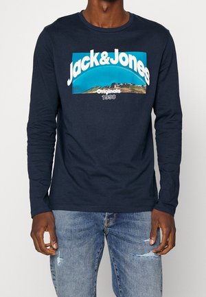 Homme portant une chemise à manches longues marine avec l'inscription "Jack & Jones Originals 1990" et un motif de montagne, associée à un jean bleu déchiré.