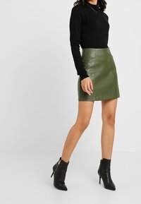 Haut noir à manches longues associé à une mini-jupe en cuir verte. Le mannequin porte des bottines noires pointues avec une fermeture éclair sur le côté.