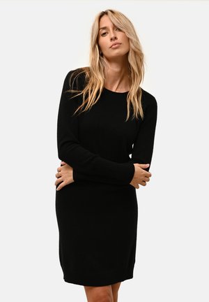 Blond kvinna som bär en svart långärmad stickad klänning står med armarna korsade mot en enkel ljus bakgrund.