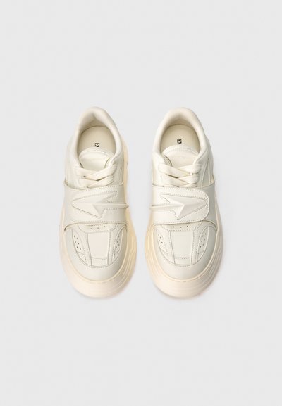 Λευκά sneakers χαμηλού προφίλ με παχιά σόλα, δέσιμο με κορδόνια και φαρδιά λωρίδα βέλκρο στο επάνω μέρος, απεικονισμένα από πάνω σε απλό φόντο.