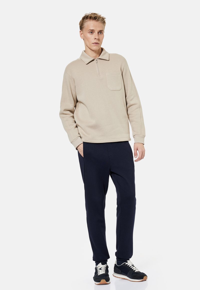 Nuori mies seisoo yhdellä kädellä taskussa, pukeutuneena vaaleanruskeaan vetoketjulliseen neuleeseen, tummiin housuihin ja mustiin tennareihin yksiväristä taustaa vasten.