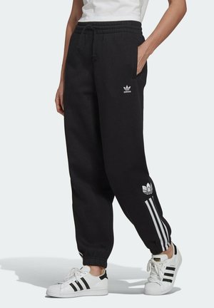 Personne portant un jogging noir Adidas avec des logos trèfle blancs et des rayures, associée à des baskets blanches Adidas avec des rayures noires.