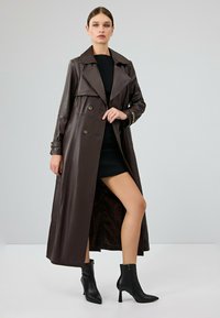 Bruin leren trenchcoat met een dubbele rij knopen aan de voorkant, oversized kraag en zijsplitten. Gemaakt over een zwarte jurk en gecombineerd met zwarte enkellaarsjes.