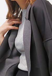 Szary sweter pod czarną, oversize'ową kurtką. Tkanina wygląda na gładką i miękką, z widocznym przeszyciem na kurtce.