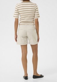 Person klädd i en beige randig kortärmad skjorta, ljusbeige shorts med rullkant och svarta ballerinas, står vänd bortifrån mot vit bakgrund.
