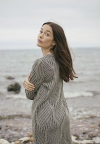 Strickkleid in Beige mit schwarzen Wellenmustern, langenÄrmeln und lockerer Passform. Der Stoff wirkt strukturiert und weich vor einem Küstenhintergrund.