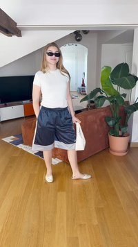 Αθλητικά μαύρα σατέν σορτς με γκρι λεπτομέρειες, σε συνδυασμό με ένα λευκό εφαρμοστό T-shirt και άνετα λευκά slip-on παπούτσια. Κρατώντας μια λευκή τσάντα.