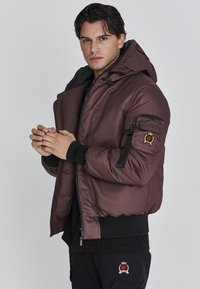 Braune isolierte Jacke mit Kapuze, Reißverschluss vorne, gerippten Bündchen und einem Logo-Patch am Arm. Das Material wirkt glatt mit einem matten Finish.