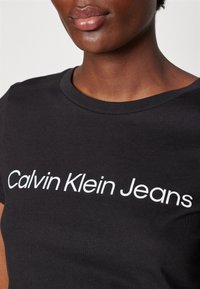 Prikaz osobe izbliza koja nosi crnu majicu s bijelim tekstom "Calvin Klein Jeans" preko prsa.
