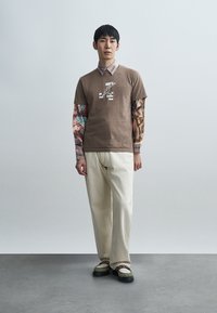 Καφέ μπλουζάκι με γραφικό τύπωμα πάνω από πουκάμισο με μακριά μανίκια και σχέδιο. Παντελόνι εκρού με φαρδιά μπατζάκια και μαύρα παπούτσια με πράσινες λεπτομέρειες.