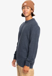 Granatowy sweter z włóczki o teksturowanej tkaninie, okrągłym dekolcie i prążkowanych mankietach. W zestawieniu z brązową czapką i khaki spodniami.