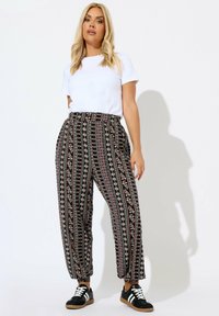 Vrouw staat in een witte t-shirt, met een patroon van hoge taille broek met bloemenstrepen en zwarte sneakers met witte strepen.