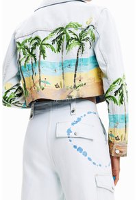 Ljus denimkort jacka med målade palmträd och strandscen, bärs med ljusa denimbyxor som har blå färgsplatter på ficka och lår.