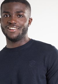 Granatowy sweter dzianinowy z okrągłym dekoltem i ściągaczami na rękawach. Na piersi znajduje się mała naszywka z logo, tkanina jest teksturowana, a krój luźny.