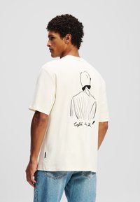 Cream short-sleeve t-shirt made of cotton, featuring a black outline illustration on the back with “Café de…” text. Relaxed fit.