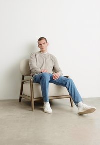 Beige genser, lyseblå jeans og hvite sneakers brukt av en mannlig figur som sitter i en lysfarget polstret stol med treben.