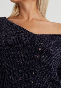 Granatowy dzianinowy sweter z odkrytymi ramionami, o plecionej fakturze i małych wycięciach zapewniających wentylację. Miękki, luźny fason.