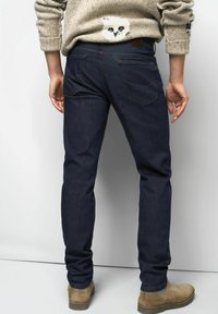 Jean en denim bleu foncé coupe slim, avec deux poches arrière et une étiquette en cuir sur la ceinture. Porté avec des bottes en daim couleur sable.