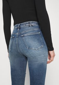 Vista trasera de una persona que lleva puestos unos jeans Calvin Klein de color azul con un lavado desgastado y una camiseta de manga larga negra, contra un fondo blanco.