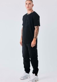 Joven con tatuajes que lleva una camiseta negra, pantalones cargo negros, zapatillas blancas, cadena de plata y anillos, de pie contra un fondo claro y liso.