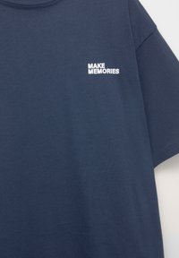 Mørkeblå t-shirt lavet af blød bomuld med korte ærmer. Har hvid broderi, der siger "SKAB MINDER" på den øverste venstre side.