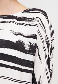 Blouse rayée noire et blanche en tissu doux et lisse. Le design comprend des motifs de coups de pinceau horizontaux de largeurs variées.