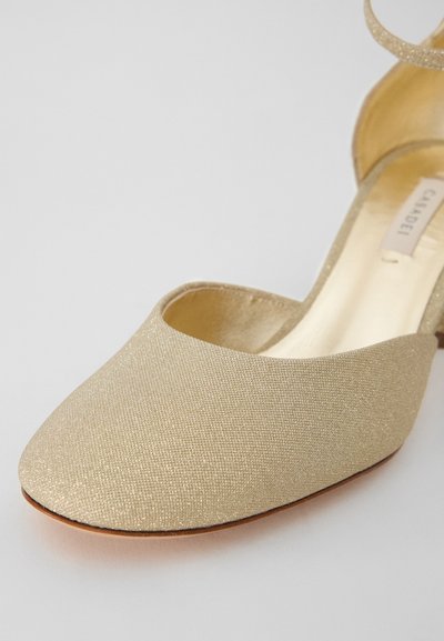 Goldfarbene, stoffgeferte Schuhe mit spitzer Zehe, glatter Textur und einem Knöchelriemen. Die Sohle besteht aus einem kontrastierenden braunen Material.