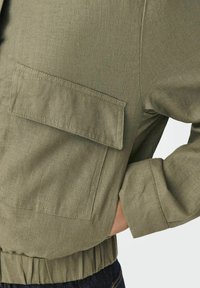 Olivovo zelená bunda z ľahkej látky, s veľkou kapsou, elastickým pásom a manžetami. Hladká textura so šitými detailmi.