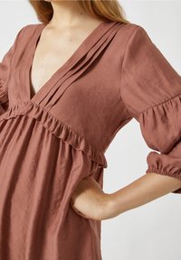 Rosenfarbenes Kleid mit tiefem V-Ausschnitt, gefaltetem Oberteil, Rüschensaum und dreiviertellangen Ärmeln. Aus weichem, strukturiertem Stoff gefertigt.