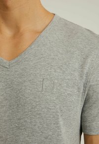 Tricou gri din bumbac, cu guler în V, featuring un logo brodat subtil în formă de pătrat pe partea stângă a pieptului. Mâneci scurte și textură moale. Design minimalist.