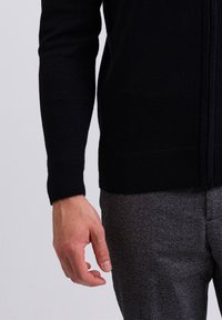 Musta neulecardigan, jossa on vetoketjukiinnitys, tekstuuripinta ja ribbireunat, yhdistetään harmaisiin kuviollisiin housuihin.