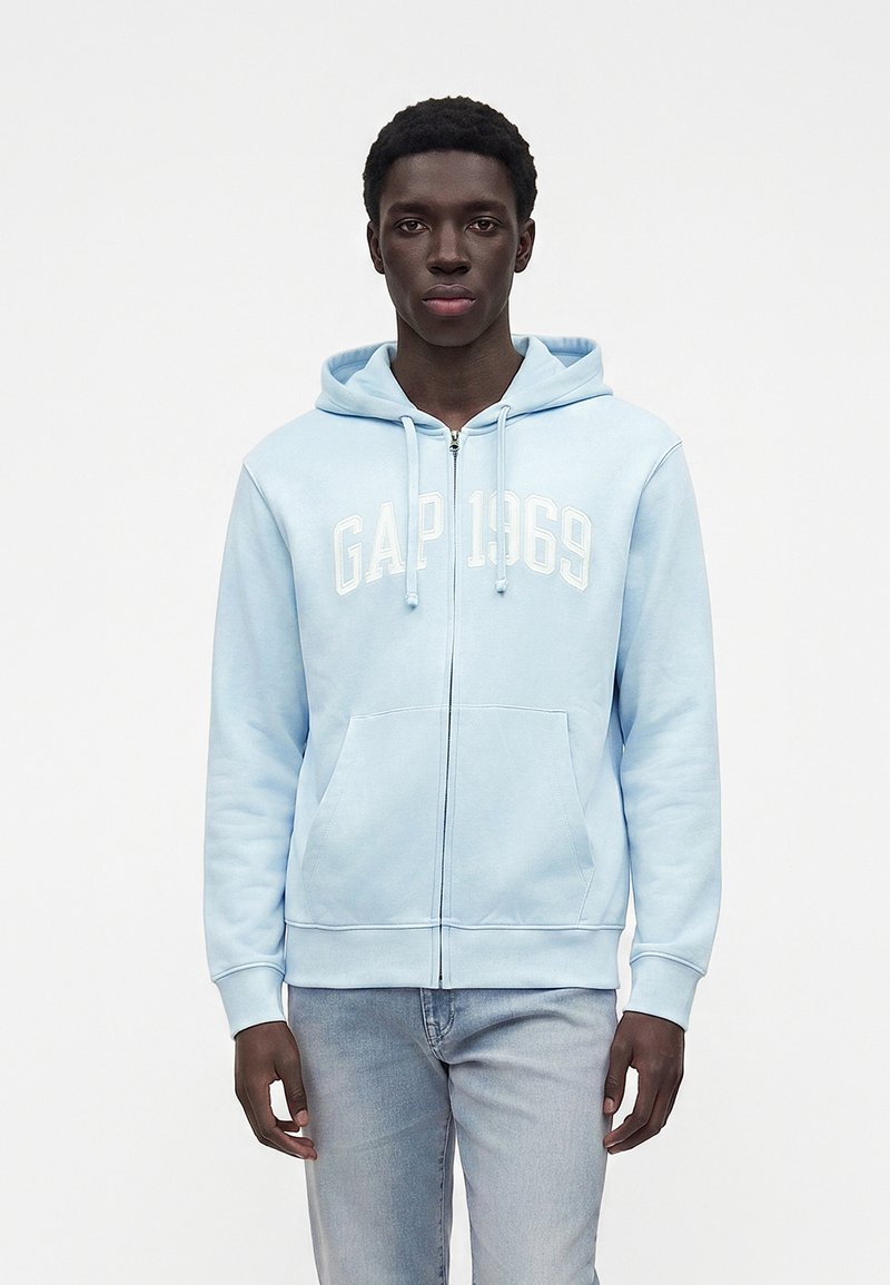 Jeune homme debout face à l’objectif, portant un sweat à capuche zippé GAP bleu clair et un jean délavé clair sur un fond blanc uni.