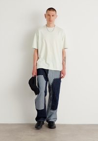 Světle zelené oversized tričko, modré a šedé džíny s patchwork vzorem a různými texturami, s předními kapsami a černými botami.