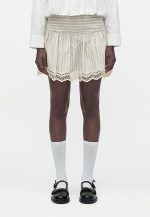 Mini-jupe rayée avec bordure en dentelle portée avec des chaussettes blanches hautes jusqu'aux genoux et des chaussures en cuir noir à boucles, associée à une chemise blanche à manches longues.