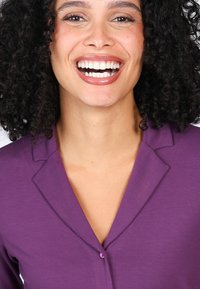 Purple collared shirt with a button closure, featuring a smooth texture and subtle sheen. Model has curly hair and a broad smile.