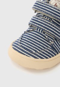 Chaussure en toile rayée bleu et blanc avec des bandes Velcro, bout arrondi et semelle en caoutchouc texturée. Présente des accents cousus et un logo.