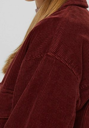 Gros plan sur une personne portant une veste en velours côtelé rouge foncé, mettant en valeur le tissu texturé et de longs cheveux blonds tombant sur l'épaule.