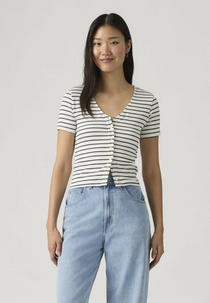 Jeune femme portant un haut boutonné à manches courtes rayé blanc et noir et un jean taille haute bleu clair, debout devant un fond neutre.