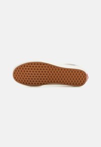 Semelle de sneaker blanche avec un motif en gaufre, en matériau en caoutchouc, couleur marron au fond et contours blancs. Présente un petit accent rouge au niveau du talon.