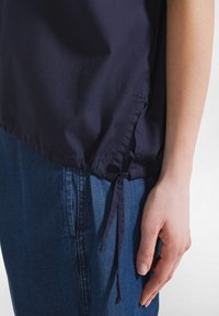 Nahaufnahme einer Hand neben blauer Jeanshose und einem marineblauen Oberteil mit gebundenem Kordelzug an der Hüfte vor weißem Hintergrund.