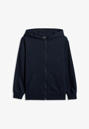 Enfant debout, les mains dans les poches, portant un bonnet rouge, un sweat à capuche zippé bleu marine, un pantalon gris et des baskets blanches aux lacets roses.
