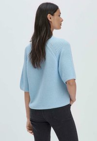 Vaaleansininen neulepusero, jossa on lyhyet hihat ja rento malli, jossa on teksturoitu kuvio ja ribbireunat, yhdistettynä tummiin farkkuihin.