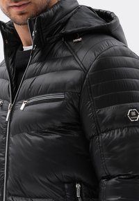 Chaqueta acolchada negra con textura de diamante, bolsillos con cremallera y capucha desmontable. Presenta herrajes plateados y un detalle de logo hexagonal.