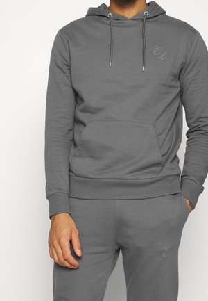 Man draagt een bijpassende grijze hoodie en joggingbroek met de initialen "CL" geborduurd op de borst en dij, staand met ontspannen handen.