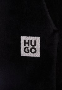 Schwarzer Stoff mit einem reflektierenden quadratischen Patch, auf dem "HUGO" in fetten schwarzen Buchstaben in zwei Reihen angeordnet ist.