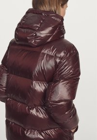 Bordeauxrode donsjas met een glanzende, doorgestikte afwerking. Voorzien van een dikke capuchon en brede horizontale strepen, die een zachte textuur benadrukken.