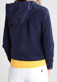 Mörkblå jacka med huva, kort längd, lätt material och knappdetaljer. Bärs över en gul skjorta och vita jeans.