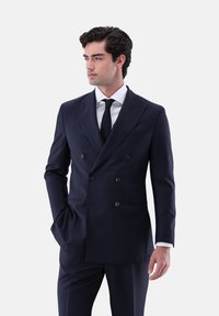 Mann in einem dunkelblauen, doppelt geknöpften Anzug mit weißem Hemd und schwarzer Krawatte, steht mit einer Hand in der Tasche und blickt nach links.