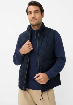 Man draagt een marineblauw doorgestikt vest over een marineblauwe trui met rits en beige broek, stelt de rits van het vest af en kijkt iets omhoog.
