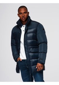 Joven vestido con una chaqueta acolchada azul marino sobre una camisa polo blanca y jeans azules, frente a un fondo claro y uniforme.